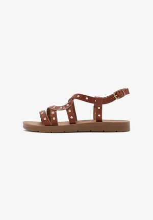 Brown leather sandals featuring multiple straps with gold studs, open sides, and an adjustable ankle buckle. Flat sole with minimal tread.