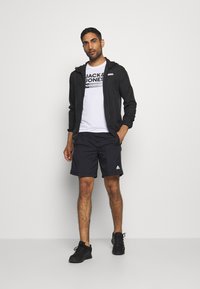 Black lightweight zip-up jacket over a white t-shirt with a bold logo, paired with black shorts and black athletic shoes.