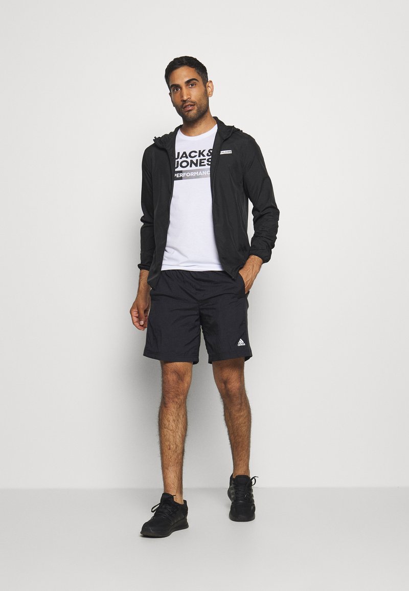 Black lightweight zip-up jacket over a white t-shirt with a bold logo, paired with black shorts and black athletic shoes.