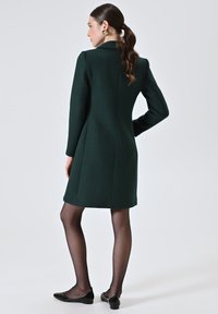Cappotto verde in lana con vestibilità su misura, maniche lunghe e taglio dritto. Indossato sopra collant neri e scarpe basse. Orecchini con dettagli dorati visibili.