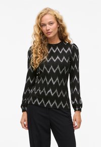 Mujer con cabello rubio rizado y largo, vistiendo una blusa negra de manga larga con patrones en zigzag plateados y pantalones negros, de pie frente a un fondo blanco.