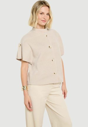 Donna con capelli biondi corti indossa un maglione beige a maniche corte abbottonato e pantaloni abbinati, in piedi con una mano rilassata di fianco.