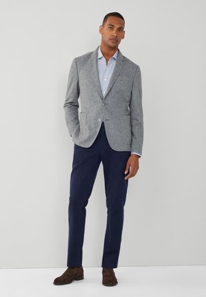 Blazer de lana gris con solapas de muesca, dos botones y bolsillos, combinado con una camisa de color azul claro con cuello y pantalones azul marino. Zapatos de ante marrón.