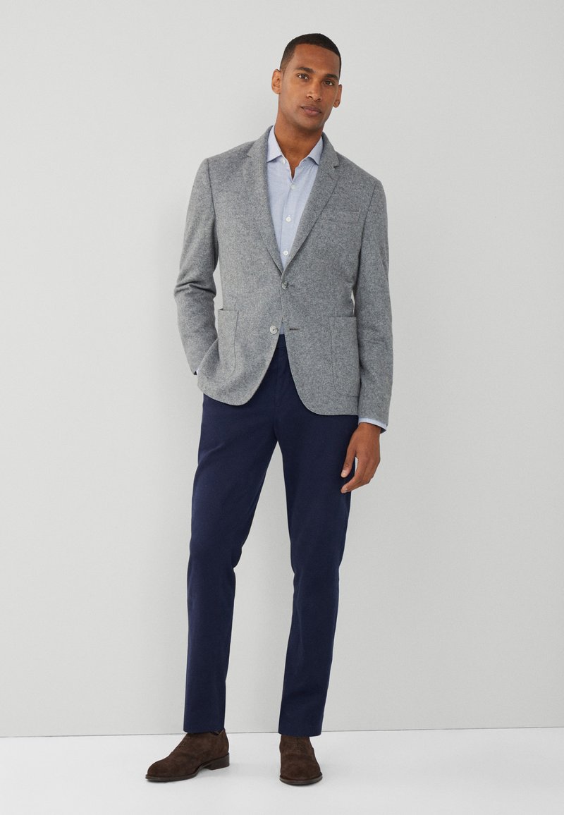 Blazer grigio in lana con revers a punta, due bottoni e tasche, abbinato a una camicia azzurra con colletto e pantaloni blu navy. Scarpe in suede marroni.