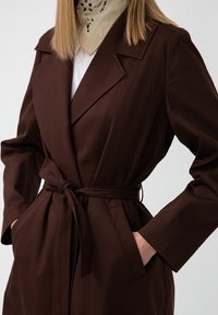 Kvinna i en mörkbrun bälte jacka med händerna i fickorna och en beige mönstrad scarf över en vit topp.