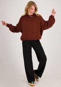 Lachende vrouw die danst, gekleed in een losse bruine trui, zwarte pantalon met krijtstreep en zwart-witte sneakers met rubberen zolen.