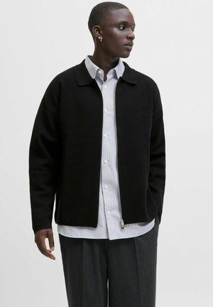 Young man with short hair wearing hoop earrings, a black zip-up jacket over a striped button-up shirt, and dark trousers, looking to the side.
