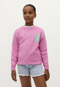 Felpa rosa a maniche lunghe con una grafica di una palma verde menta nella zona della tasca anteriore. Abbinata a pantaloni corti di jeans chiari.