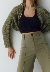 Olijfgroen opengewerkt vest over een zwarte aansluitende tanktop, gecombineerd met hooggepaste, licht olijfgroene wijde broek met verticale naden.