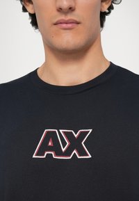 Camiseta de algodón negra con un destacado logo "AX" en rojo y blanco en el pecho. Diseño de cuello redondo, textura suave.