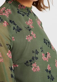 Grüne Bluse mit pinkem und schwarzem Blumenmuster sowie einem gerafften hohen Halsausschnitt, getragen von einer Person mit braunen Haaren.