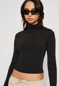 Langermet svart turtleneck topp laget av glatt materiale, med en tettsittende design og avkortet kant. Brukt med svarte solbriller.