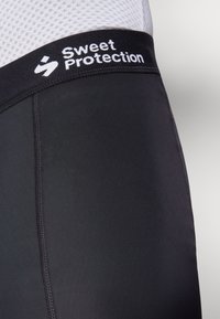 Črne športne kratke hlače z gladko teksturo, ki imajo kontrastni pas z napisom "Sweet Protection." Šivni detajli ob straneh.