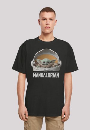 Camiseta de algodón negra con un gráfico de un personaje verde en una cápsula, enmarcado por el texto "THE MANDALORIAN" en letras blancas.