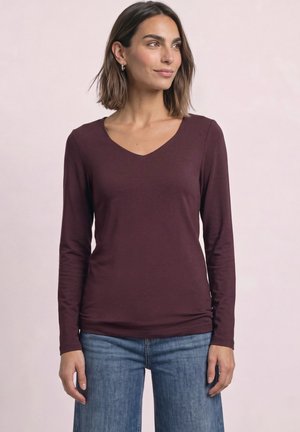 Kvinde iført en langærmet mørkelilla V-hals trøje og blå jeans, stående mod en ensfarvet lys baggrund og ser let mod højre.