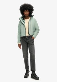 Lichtgroene cropped jas met een capuchon, zwarte accenten, over een crèmekleurige trui, gecombineerd met hoog-waisted grijze jeans en zwarte veterschoenen.