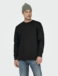 Schwarzes Langarm-T-Shirt aus Baumwolle, mit Rundhalsausschnitt und lockerer Passform. Kombiniert mit hellblauen Jeans und einer grauen Strickmütze.