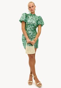 Vestido verde de lentejuelas con mangas abullonadas, diseño ajustado, combinado con un bolso de paja. Sandalias de plataforma doradas y joyería a juego completan el conjunto.