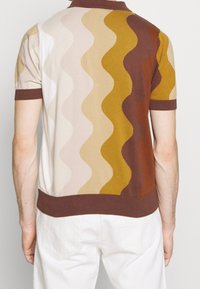 Muž v tričku s krátkým rukávem pleteným s vertikálními vlnitými pruhy v krémové, béžové, hořčicové, hnědé a rezavé barvě, spárovaném s bílými kalhotami.