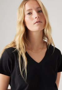 Tricou negru cu decolteu în V și mâneci scurte, fabricat dintr-un material neted. Modelul are părul ondulat și blond și poartă un colier subțire din aur.