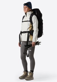 Vit och beige vattentät jacka, grå leggings och bruna vandringskängor. Svart ryggsäck som håller vandringsstavar, med en blå mössa.