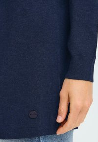 Pull en tricot bleu marine avec des poignets côtelés, mettant en avant un détail de bouton visible à l'ourlet. Texture lisse et coupe décontractée.