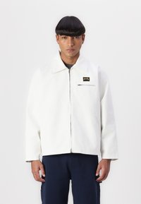 White zip-up jacket with a collar, featuring a black chest pocket and a logo patch. Smooth texture and relaxed fit.