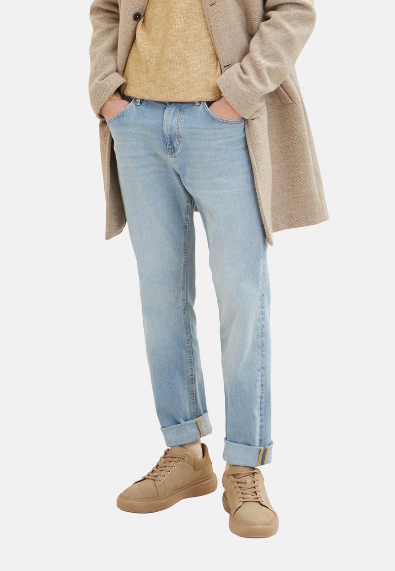 Blugi din denim de un albastru deschis, cu un croi lejer, manșete răsucite și accente galbene. Asortați cu adidași bej și un pulover din tricot bej sub o haină.