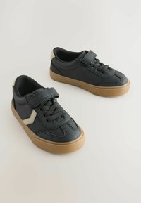 Baskets noires avec une sangle velcro et des lacets, présentant un accent beige plus clair et une semelle en caoutchouc, exposées sur un fond neutre.