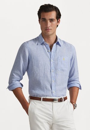 Hellblau gestreiftes Hemd aus Baumwolle mit einer Brusttasche und hochgekrempelten Ärmeln, kombiniert mit weißen Hosen und einem braunen Gürtel.