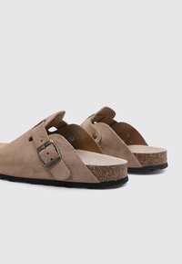 Braune Wildledersandalen mit einem Korkfußbett, verstellbarer Metall-Schnalle und strukturiertem Gummisohle; offener Rücken und seitliche Aussparungen.