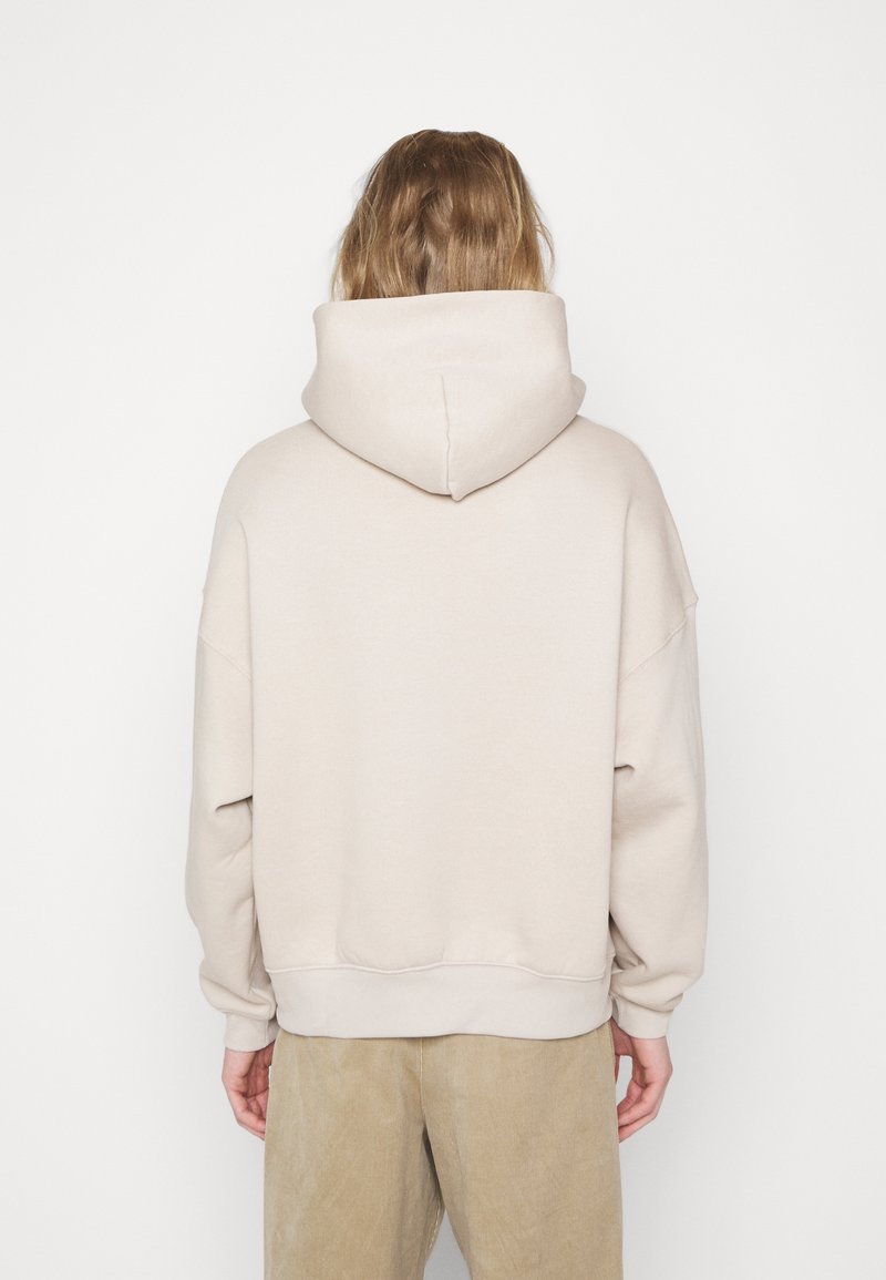 Personne aux cheveux mi-longs, vue de dos, portant un sweat à capuche beige uni et un pantalon marron clair sur fond blanc.