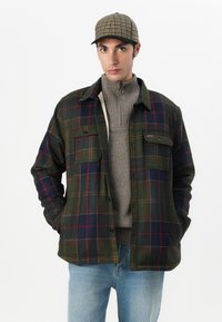Karierte Flanelljacke in Dunkelgrün, Blau und Rot. Zwei Brusttaschen, geknöpfte Vorderseite, über einem grauen gerippten Rollkragenpullover getragen. Modell trägt eine Mütze.