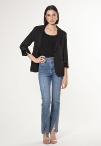 Blazer nero con rifinitura in paillettes sulle maniche, abbinato a jeans in denim blu a vita alta con spacchi frontali. La modella indossa sandali argentati.