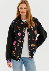 Czarna bomberka w kwiatowy wzór z kolorowymi motywami, z zatrzaskami i różowymi kieszeniami. Ma luźny krój i jest wykonana z miękkiego materiału.