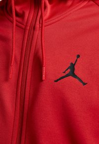 Sudadera roja con cremallera fabricada en tela suave, que presenta el logo negro de Jumpman en el pecho, con cordones texturizados y un ajuste casual.