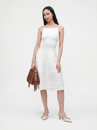 Femme portant une robe blanche sans manches au genou, tenant un sac à main marron et portant des chaussures blanches à talons bas sur un fond uni.