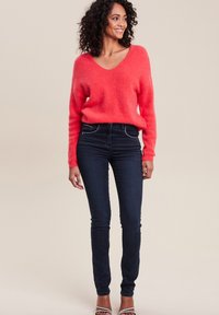 Pull v-neck en corail duveteux à manches longues associé à un jean skinny foncé. Le modèle se tient debout, mettant en valeur une coupe décontractée et une texture douce.