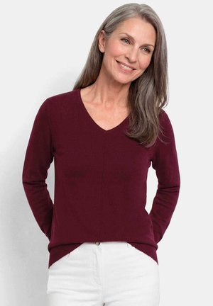 Glunderende volwassen vrouw met lang grijs haar, gekleed in een bordeauxrode V-hals trui en witte broek, staand tegen een effen achtergrond.