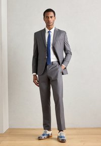 Grå dress med snittlapeller, hvit skjorte, blå slips og mønstrede dressko i blå og grå toner, satt mot en lys bakgrunn.