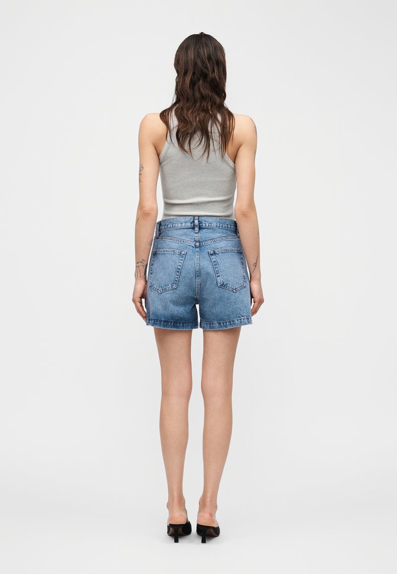 Frau mit langen braunen Haaren, die ein graues Tanktop, blaue Jeansshorts und schwarze High Heels trägt, steht mit dem Rücken zur Kamera.