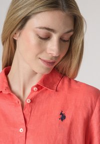 Camicia button-up in lino corallo con colletto classico e logo ricamato sul lato sinistro del petto. Texture morbida con una sensazione leggera.