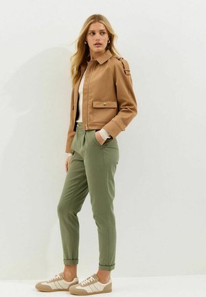 Jeune femme portant une veste courte marron clair, un haut blanc, un pantalon vert olive et des baskets beige, debout devant un fond clair uni.