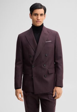 Mann trägt einen dunkelvioletten, zweireihigen Anzug und einen schwarzen Rollkragenpullover und steht vor einem hellgrauen Hintergrund.