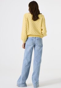 Vrouw met lang bruin haar, gekleed in een gele gebreide trui en lichte, wijde jeans, staand met haar rug naar de camera tegen een effen achtergrond.