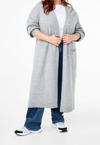 Ljusgrå lång cardigan i mjukt tyg, med två frampockets och öppen framdesign, lager-på-lager över en vit topp och blå jeans.