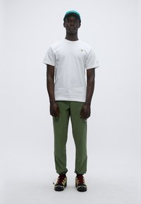 Λευκό κοντομάνικο μπλουζάκι, πράσινο παντελόνι και μαύρα παπούτσια με κόκκινες λεπτομέρειες. Το μπλουζάκι έχει ένα μικρό λογότυπο· το παντελόνι διαθέτει διακριτικές ραφές.