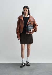 Rjava usnjena krajšana jakna, črna pulover, rjava krilo, bele nogavice, črna torbica in črne čevlje z belimi detajli. Gladke teksture in plastenjsko oblikovanje.