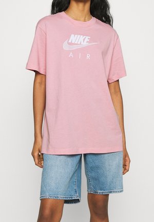 Personne portant un t-shirt Nike Air rose ample et un short en jean bleu clair, debout sur un fond blanc uni.