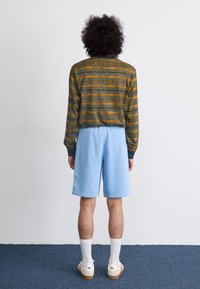 Langermet, grønn og sennepsfarget stripete topp med grå mansjetter, kombinert med lyseblå shorts. Modellen står på en blå teppebakgrunn.